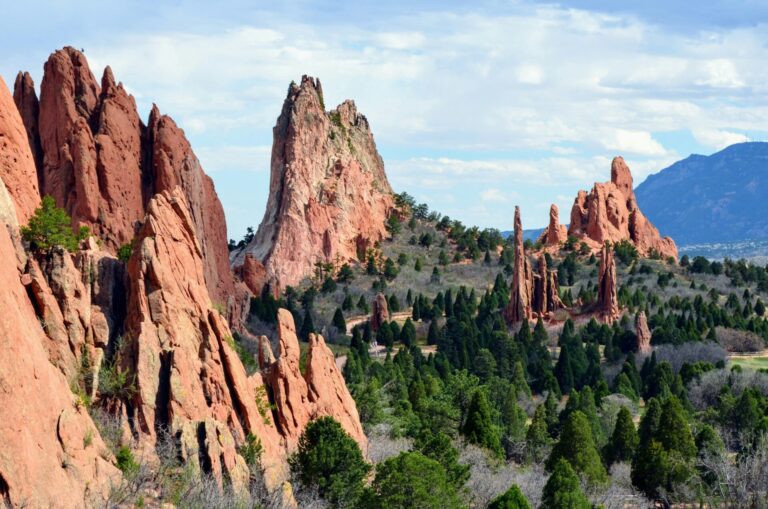 Red rock formations rise among green pines under a cloudy blue sky near Colorado Springs, CO.