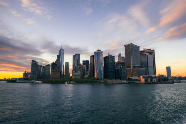 New York City skyline at sunset, with skyscrapers reflected in harbor waters.