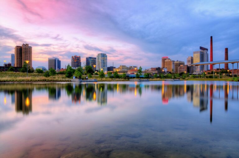 Birmingham, AL, sits beneath a colorful sky, with a body of water reflecting the skyline.