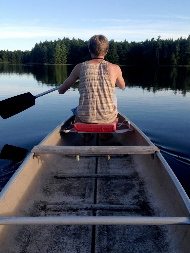 A man sitting on a red padded cushion in a canoe paddles across a calm lake, with a forest of trees on the other side.