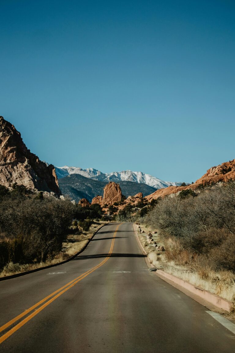 Empty road leading through Garden of the Gods, with red rock formations and snow-capped mountains in the distance under clear blue sky.