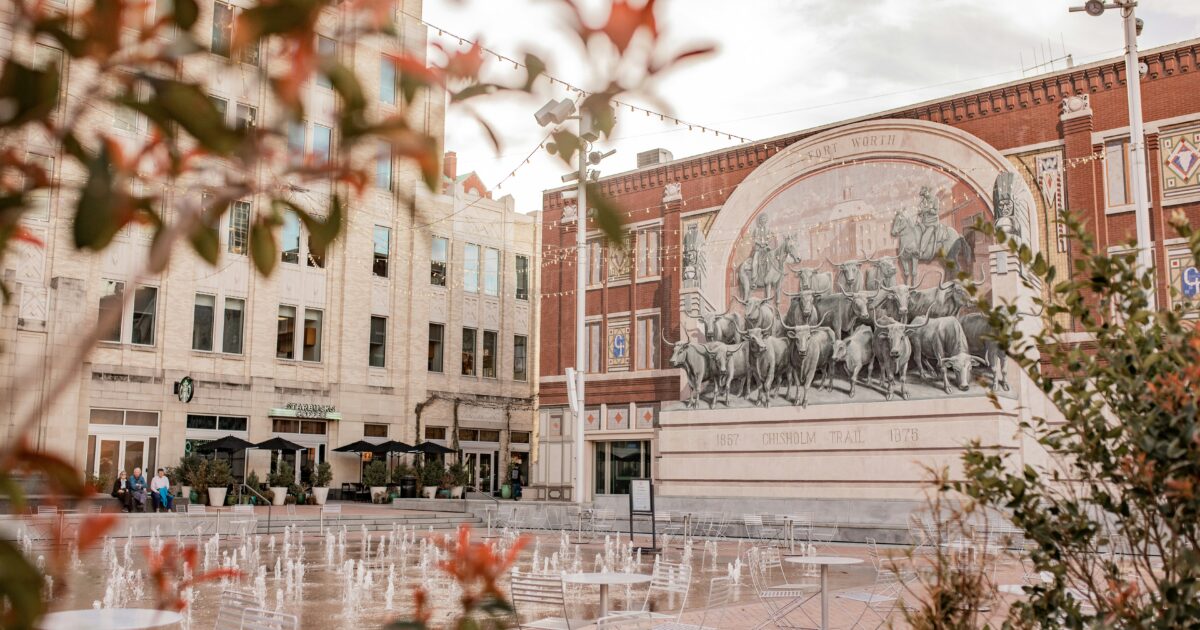 A building with a mural of cattle behind an open area with tables and chairs and several small fountains.