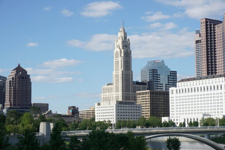 Columbus, Ohio skyline featuring the distinctive LeVeque Tower and other skyscrapers, with the Scioto River and a bridge in the foreground.