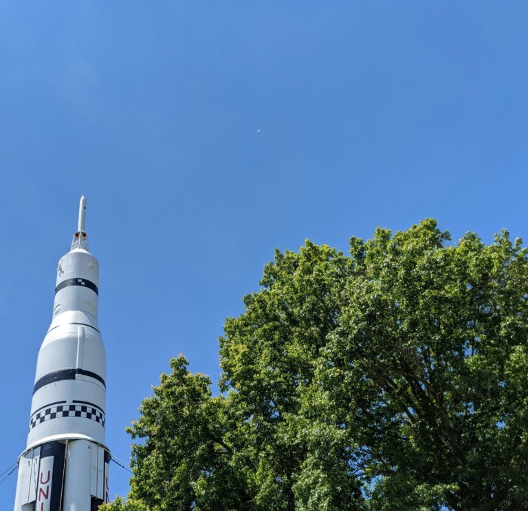 Aerial view of a large green, full treetop beside a white-and-black rocket against a clear blue sky.