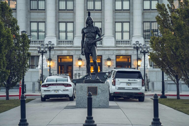 Statue of Native American figure in front of government building, flanked by two vehicles and ornate lampposts.