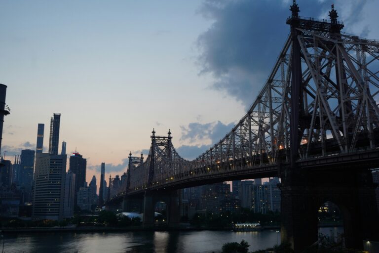 A cantilever bridge spans a river with the Manhattan skyline on the far side and the Queens waterfront in the foreground.