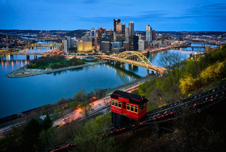 The Duquesne Incline descends a hillside in Pittsburgh at dusk, with the city's skyline, bridges, and rivers illuminated in the background.