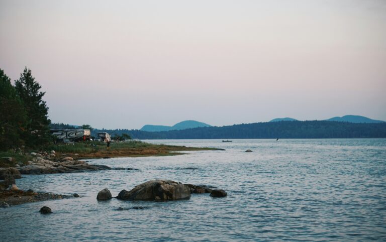 Body of water surrounded by trees with an RV and hills in the distance.