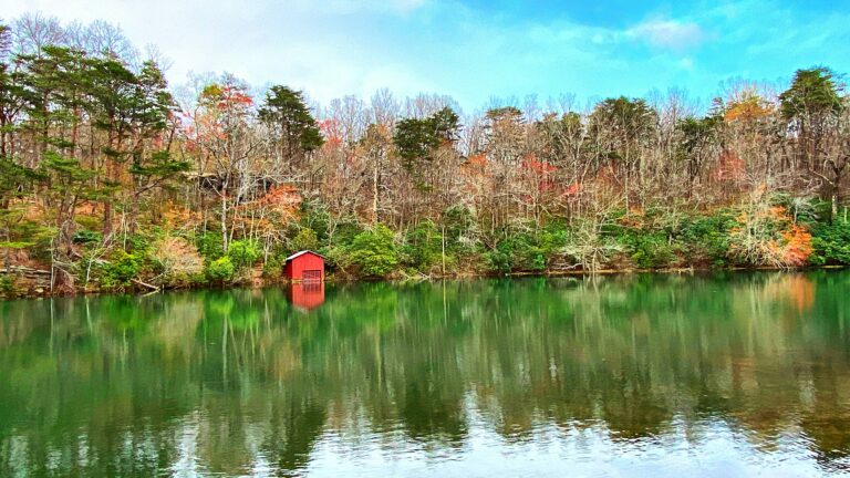 A small red boathouse sits by a calm lake, surrounded by autumn trees.