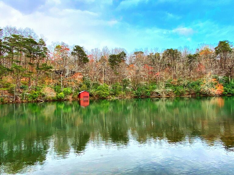 A small red boathouse sits by a calm lake, surrounded by autumn trees.