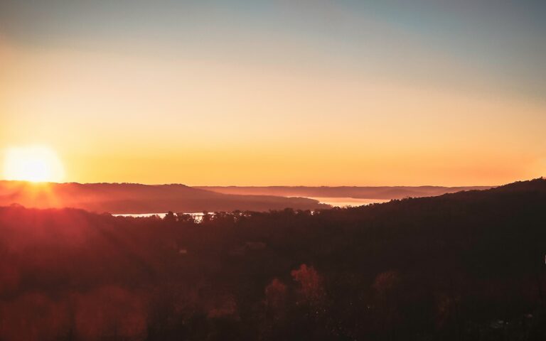 A golden-red autumn sunset over oak and maple trees with a winding lake in the background.