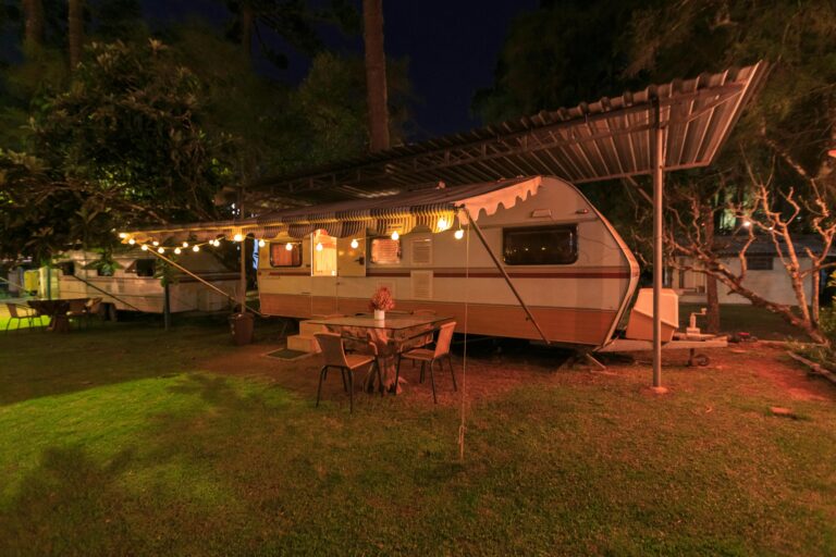 A camper parked under a wooden deck near cypress trees with a festive table and chairs under a lighted canopy outside.