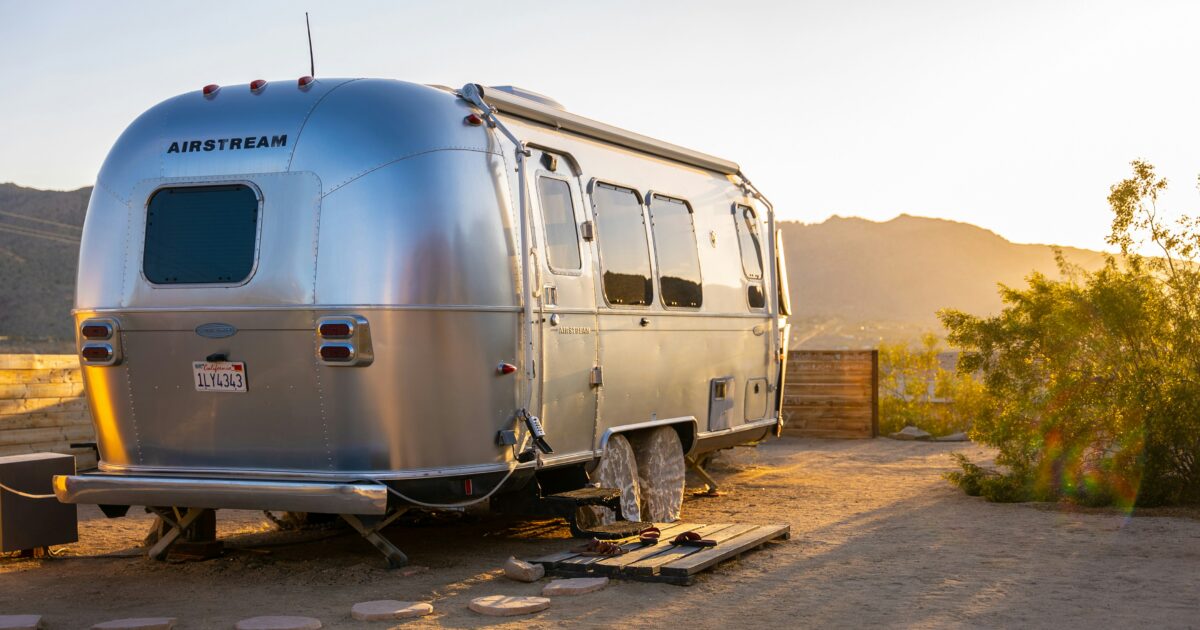A shining silver travel trailer parked on a patch of dirt with hills in the background under a sunlit sky.