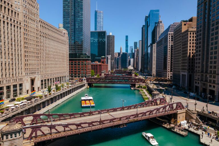 Two boats float along a bridge-covered river flanked by skyscrapers on a bright day under a blue sky