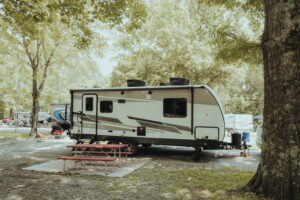 This image is a photograph of a campsite with a travel trailer and a group of people sitting outside underneath trees.