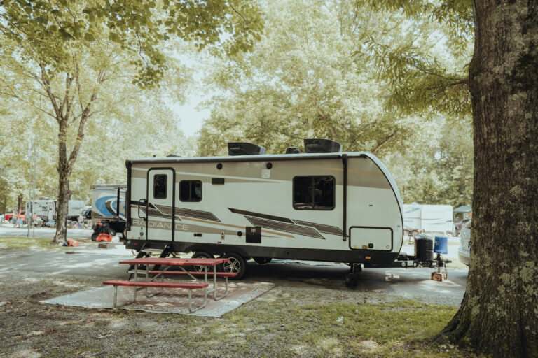 This image is a photograph of a campsite with a travel trailer and a group of people sitting outside underneath trees.
