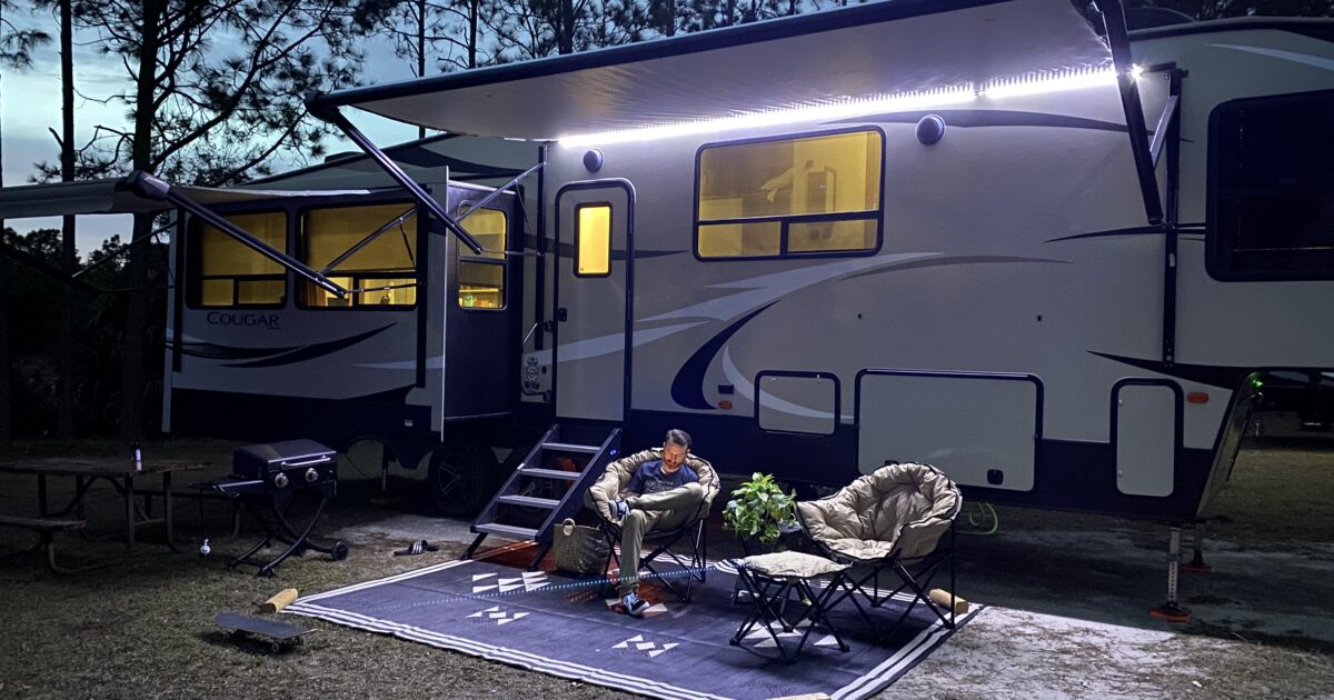 A man relaxes outside of his RV under a lit canopy