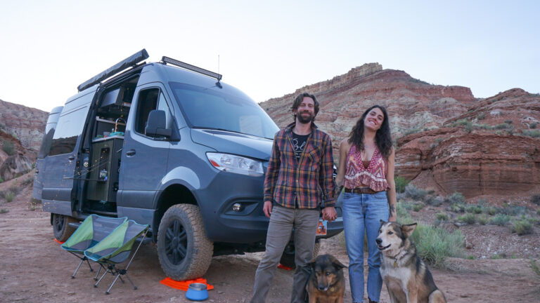 man and woman standing in front of their campervan with two dogs and red rocks in the background