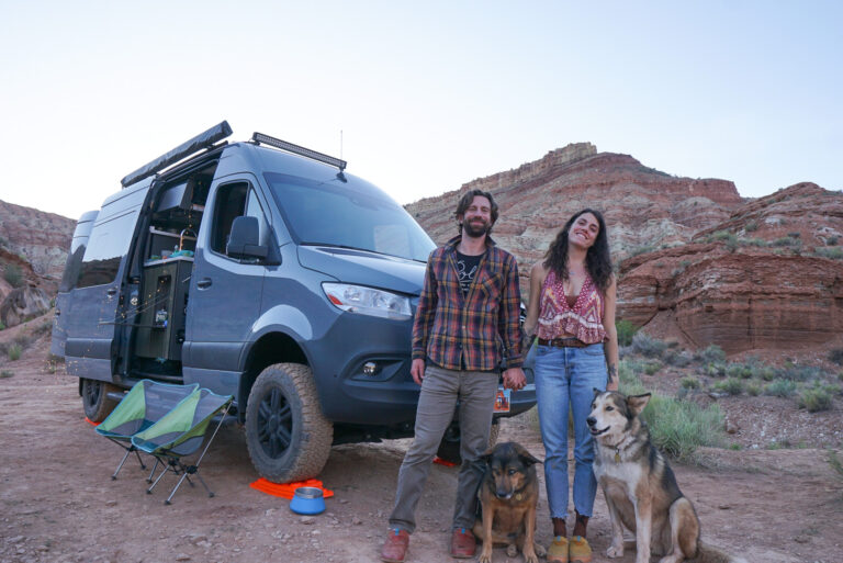 man and woman standing in front of their campervan with two dogs and red rocks in the background
