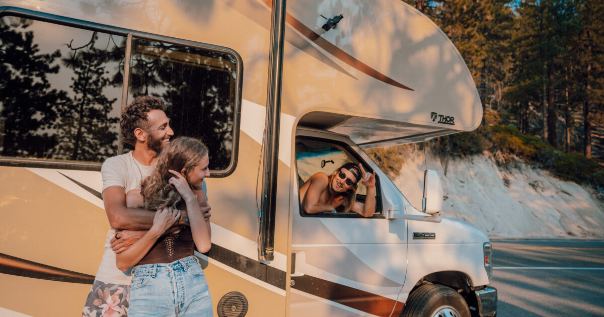 A couple hugs in front of an RV while their friend smiles out the passnger-side window