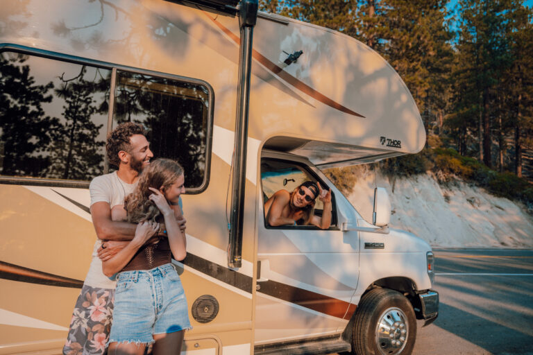 A couple hugs in front of an RV while their friend smiles out the passnger-side window