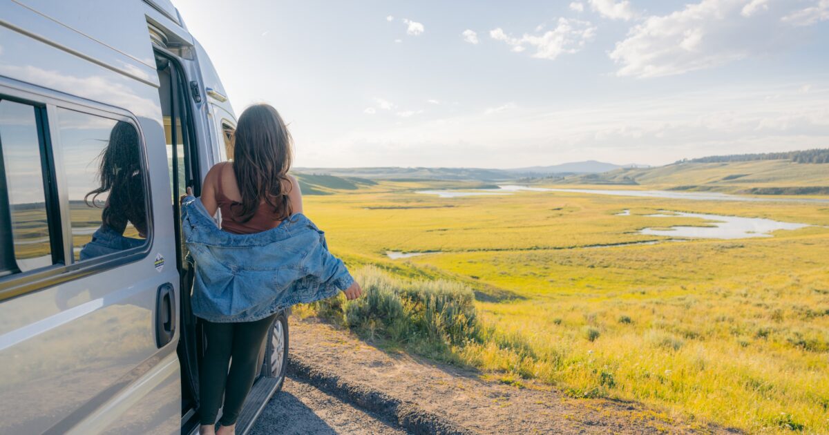 A woman leans out of her Class B campervan in front of a field of grass and mountains