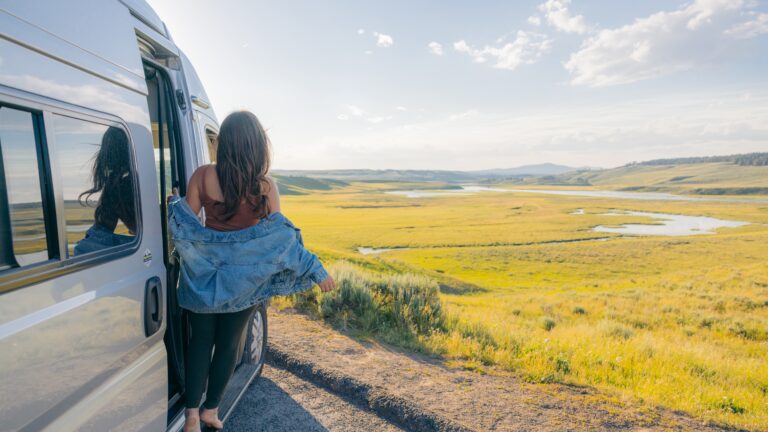 A woman leans out of her Class B campervan in front of a field of grass and mountains