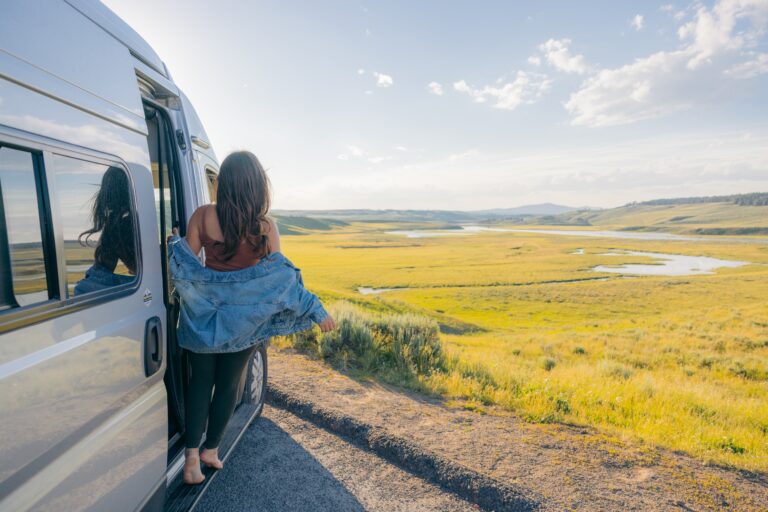 A woman leans out of her Class B campervan in front of a field of grass and mountains
