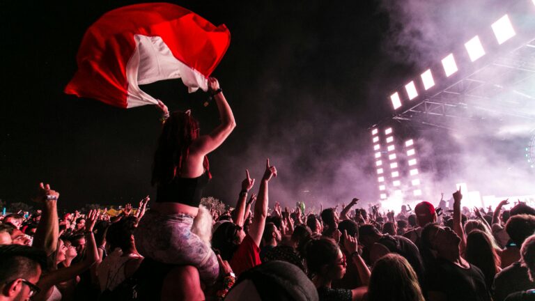 A young woman sits atop her companion's shoulders, cheering at a nightime show at a music festival