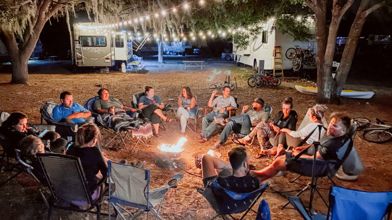 A large group sits around a campfire between two RVs with fairy lights strung between them