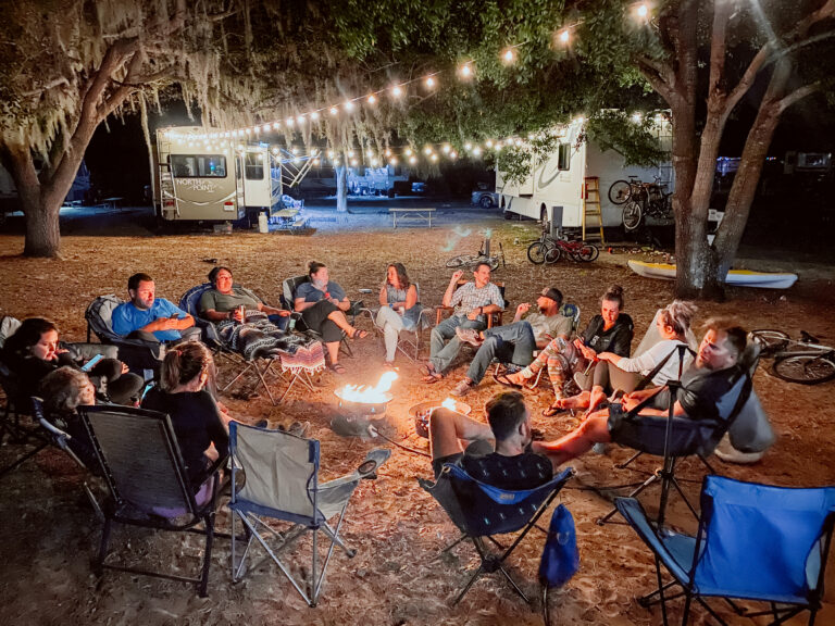 A large group sits around a campfire between two RVs with fairy lights strung between them
