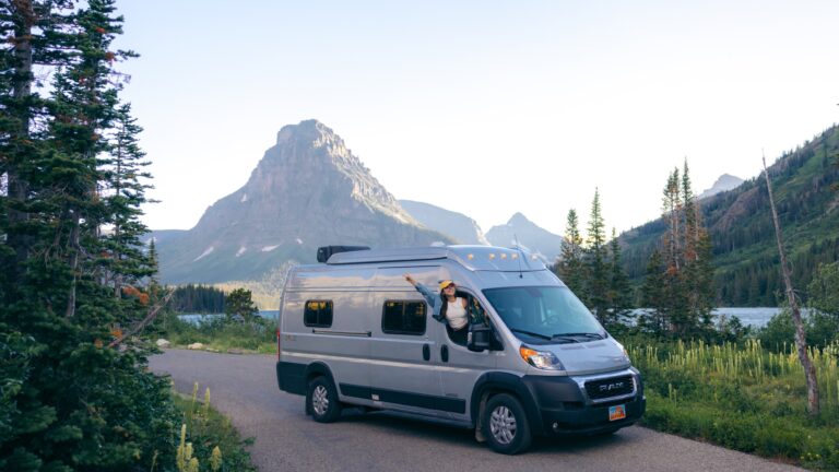 A woman leans out of a Class B van in front of a beautiful mountain landscape