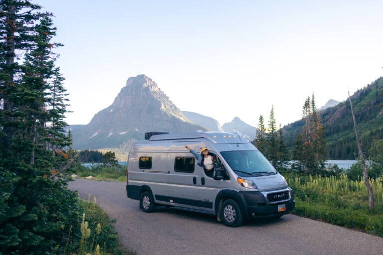 A woman leans out of a Class B van in front of a beautiful mountain landscape