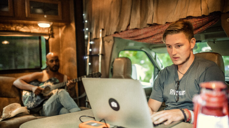 A man works on a laptop inside an RV while his friend sits on the couch playing guitar