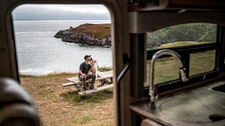 A man and his dog sit outside of an open RV door in front of a beautiful waterside area