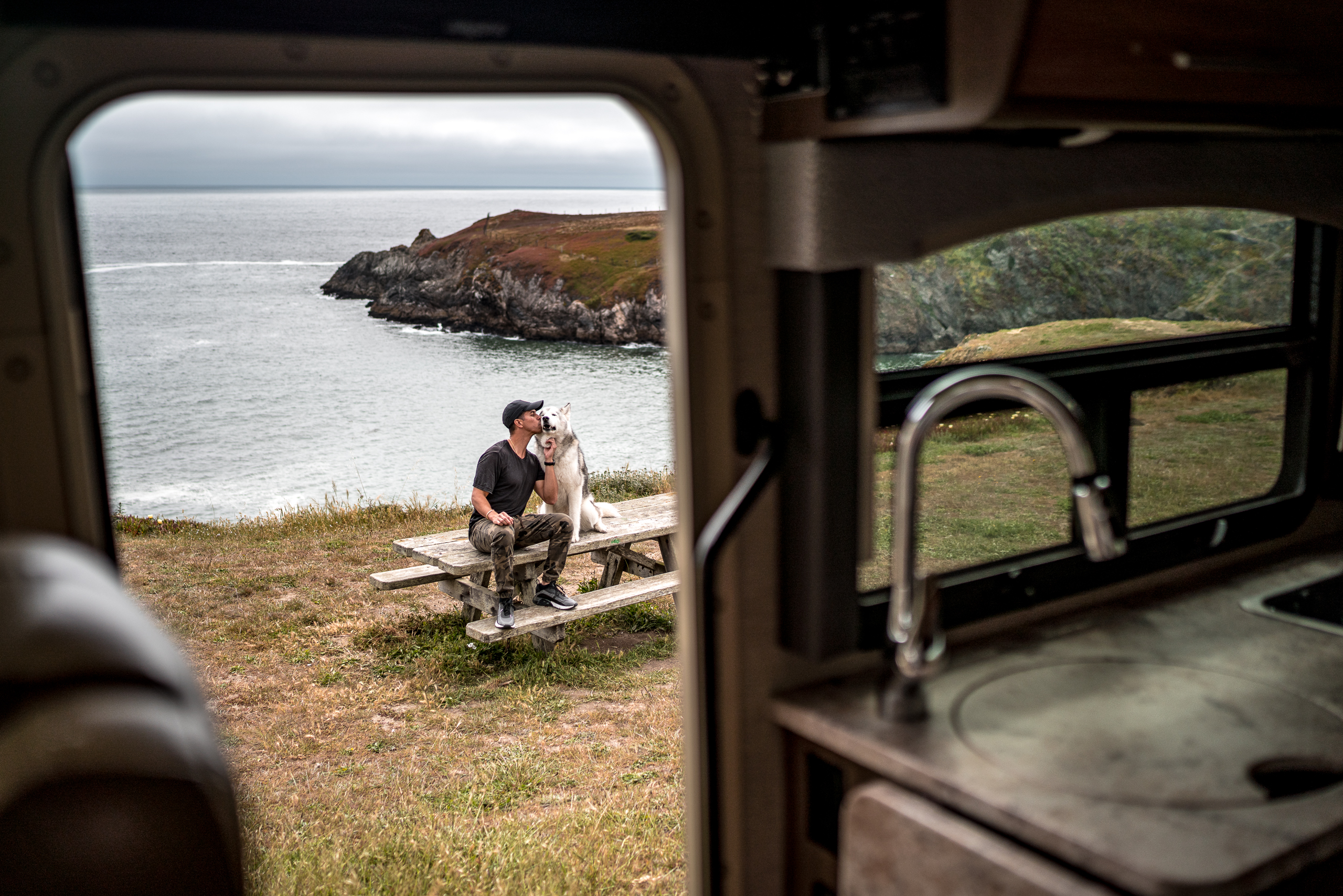 A man and his dog sit outside of an open RV door in front of a beautiful waterside area