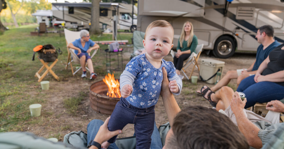 A man holds up a baby in front of a campfire surrounded by people in front of a motorhome