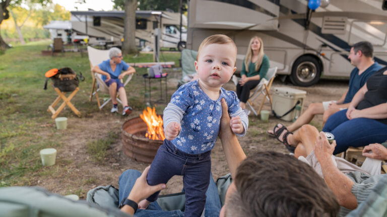 A man holds up a baby in front of a campfire surrounded by people in front of a motorhome