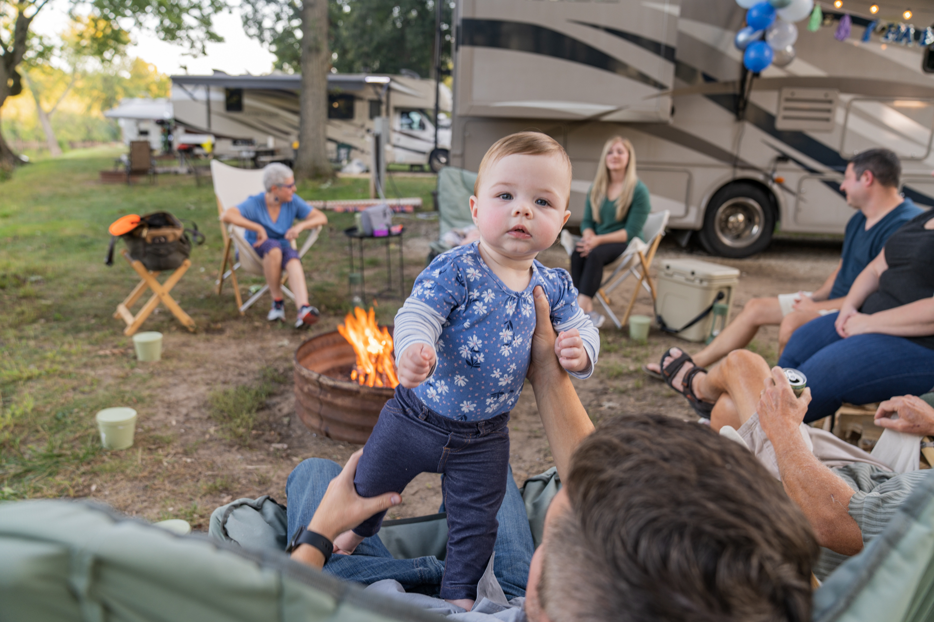 A man holds up a baby in front of a campfire surrounded by people in front of a motorhome