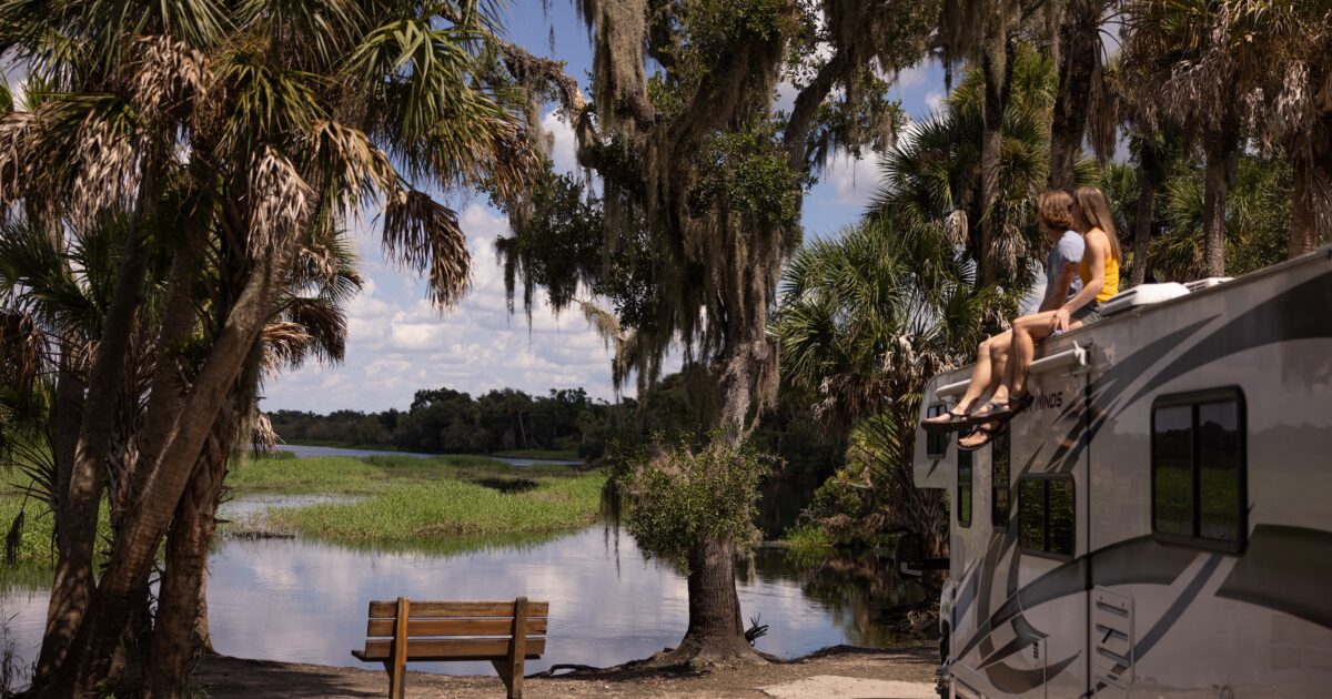 A family sits on the roof of an RV near a beautiful waterfront with palm trees