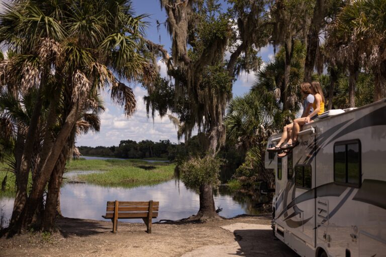 A family sits on the roof of an RV near a beautiful waterfront with palm trees