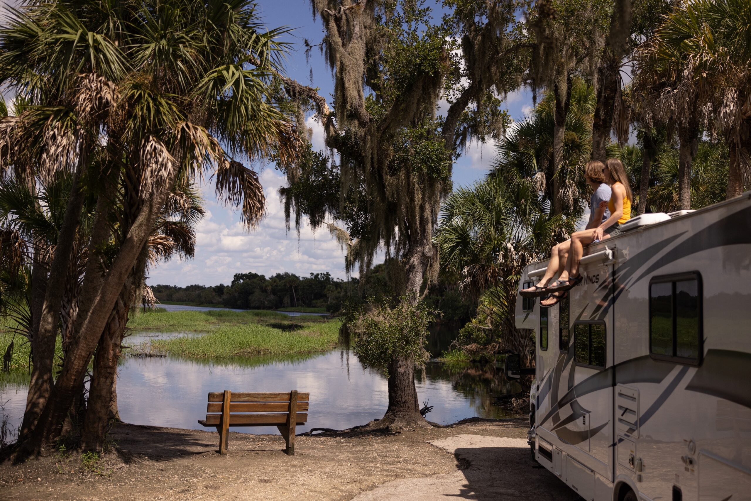 A family sits on the roof of an RV near a beautiful waterfront with palm trees